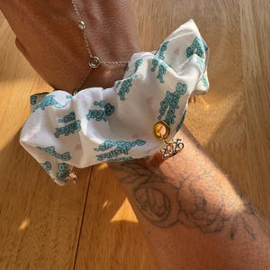 May include: A white scrunchie with a teal "Backstreet Boys" print and a gold charm with the number "2026" hangs from a silver hand chain. The scrunchie is attached to a wrist.