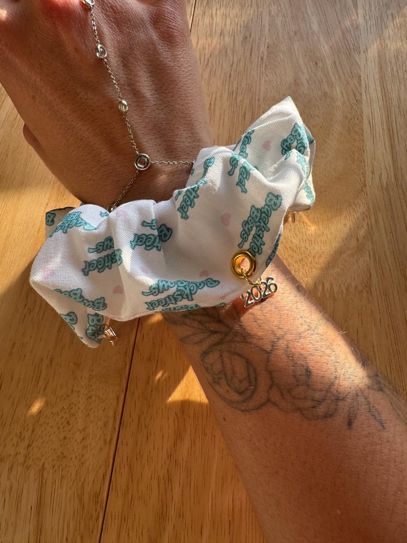 May include: A white scrunchie with a teal "Backstreet Boys" print and a gold charm with the number "2026" hangs from a silver hand chain. The scrunchie is attached to a wrist.