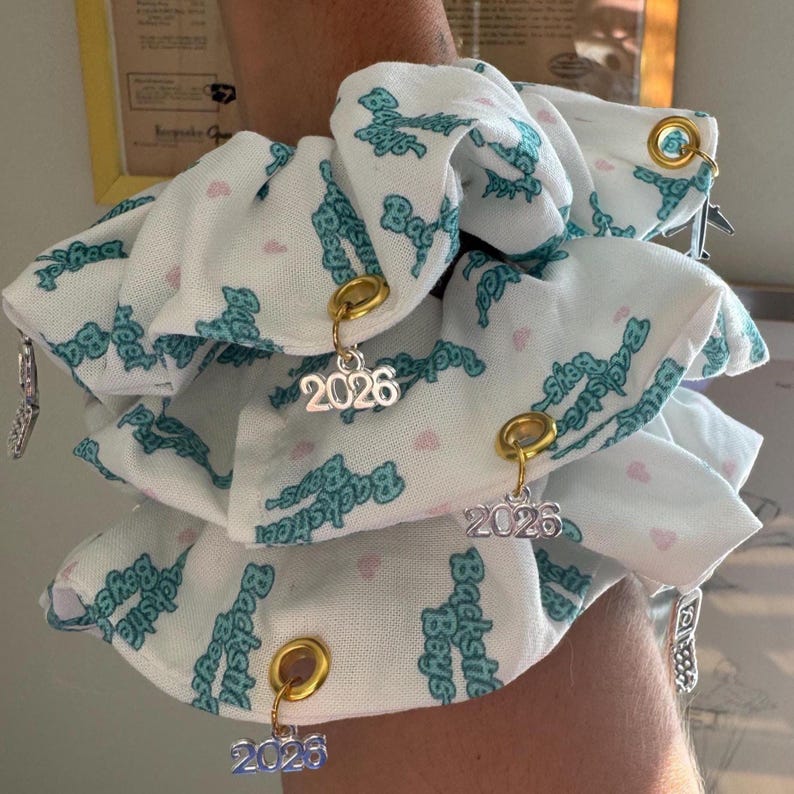 May include: White scrunchies with a repeating teal "Backstreet Boys" pattern and pink hearts. Each scrunchie has a gold or silver charm with the number "2026" hanging from it. The scrunchies are accessorized with small metal charms.