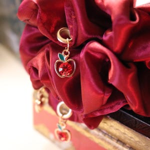 May include: A burgundy scrunchie with gold apple charms. Each charm features a red gemstone center, a gold outline, and a green leaf. The scrunchie is made of a soft, shiny fabric.