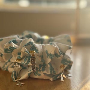 May include: White scrunchies with a teal pattern and gold-colored charms. Charms include a flip phone, an airplane, and a crown. The scrunchies are made of a soft fabric and are gathered to create a voluminous look.