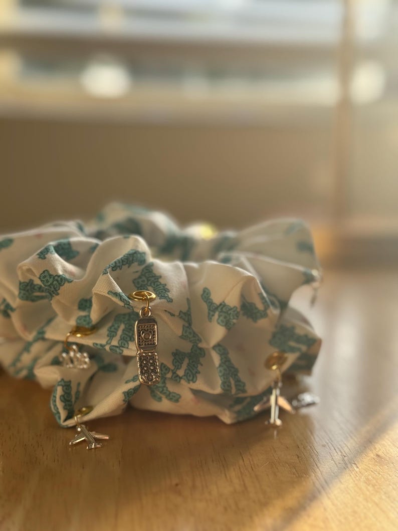 May include: White scrunchies with a teal pattern and gold-colored charms. Charms include a flip phone, an airplane, and a crown. The scrunchies are made of a soft fabric and are gathered to create a voluminous look.