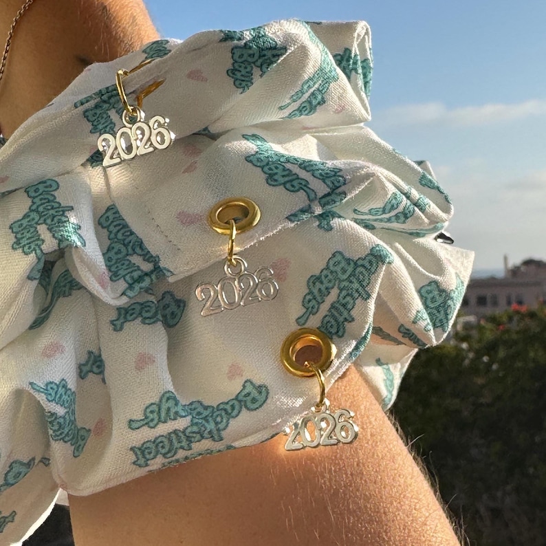 May include: Close-up of a white fabric with a teal pattern and gold-colored grommets. Three silver charms with the number "2026" are attached to the fabric. The fabric has a repeating pattern of the words "Backstreet Boys".