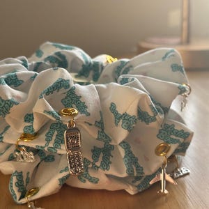 May include: A white scrunchie with a teal pattern and gold and silver charms. Charms include a flip phone, airplane, and other small shapes. The scrunchie is made of a soft fabric and is gathered.