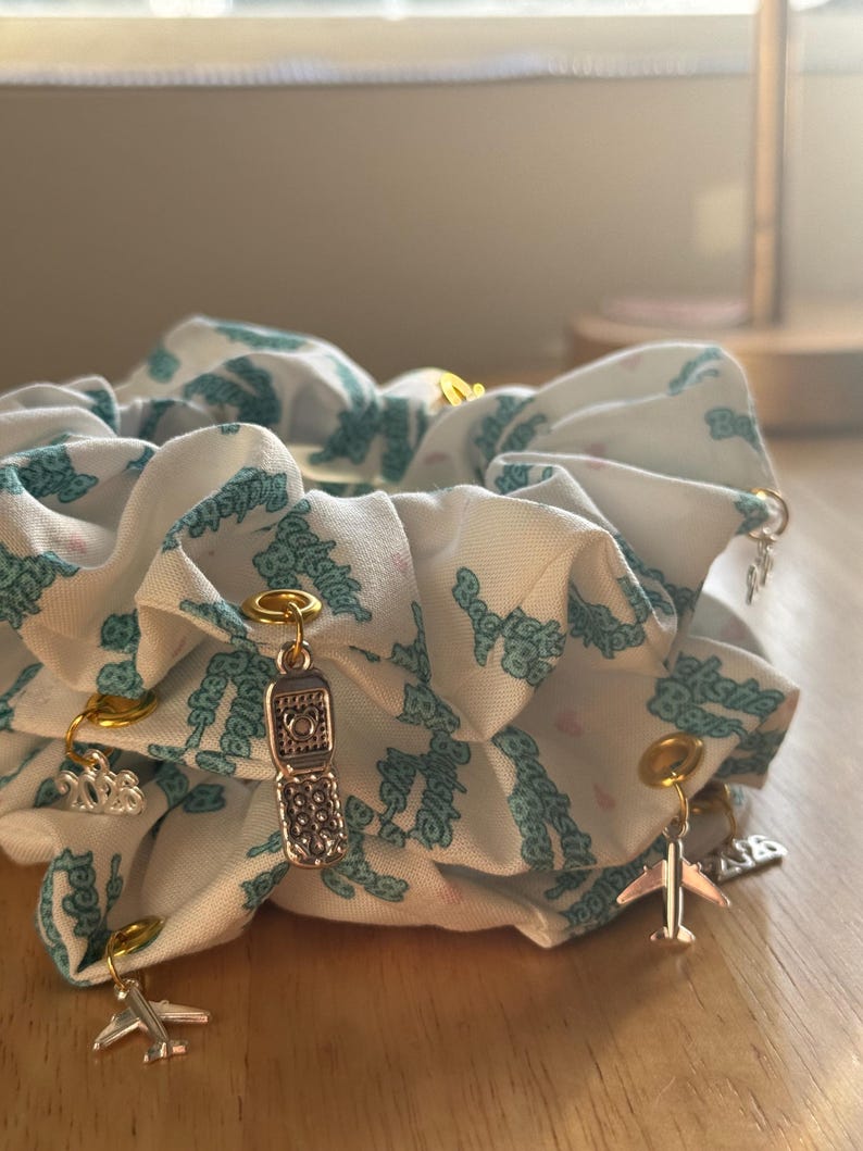 May include: A white scrunchie with a teal pattern and gold and silver charms. Charms include a flip phone, airplane, and other small shapes. The scrunchie is made of a soft fabric and is gathered.