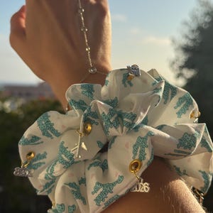 May include: White scrunchie with a repeating teal pattern and gold-colored charms. Charms include a small airplane, the number "2026", and other decorative elements. A hand chain and rings are also visible.