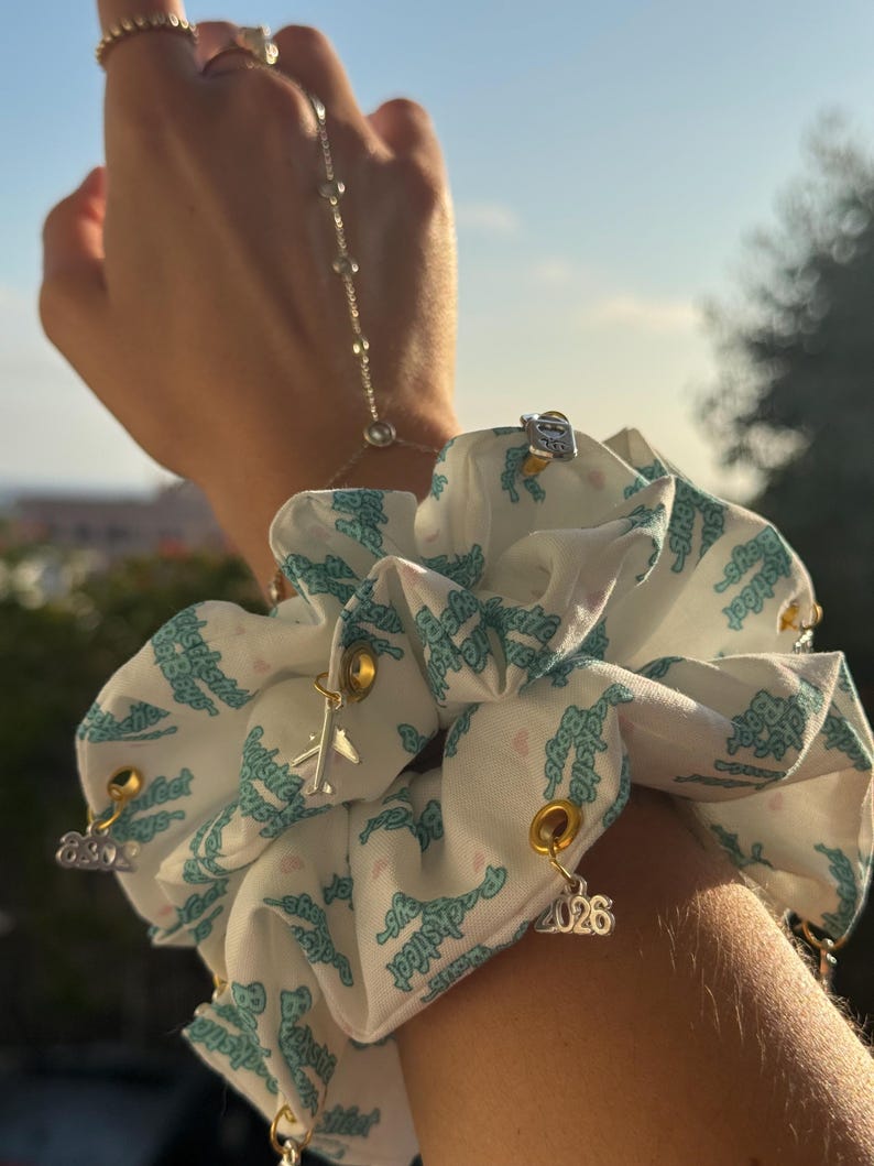 May include: White scrunchie with a repeating teal pattern and gold-colored charms. Charms include a small airplane, the number "2026", and other decorative elements. A hand chain and rings are also visible.