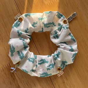 May include: White scrunchie with a repeating teal "Backstreet Boys" text pattern and small pink hearts. Adorned with gold charms featuring the numbers "2023" and "2026", a silver charm, and a small airplane charm.