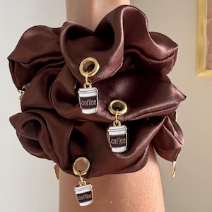 May include: A brown satin scrunchie with three gold-toned charms. Each charm features a white coffee cup with the word "coffee" in black. The scrunchie is a rich, dark brown color.