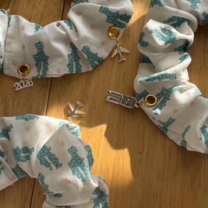 May include: White scrunchies with a teal "Backstreet Boys" pattern and small pink hearts. Each scrunchie has a gold charm, including a plane, a ticket, and the number "2026". These accessories are perfect for hair styling.