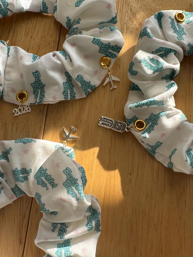May include: White scrunchies with a teal "Backstreet Boys" pattern and small pink hearts. Each scrunchie has a gold charm, including a plane, a ticket, and the number "2026". These accessories are perfect for hair styling.