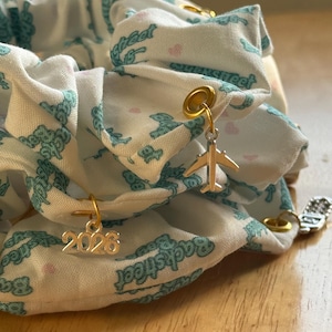 May include: A white scrunchie with a teal pattern and gold charms. The scrunchie has a repeating pattern of teal teddy bears and the word "Backstreet". Gold charms include a small airplane and the number "2026".