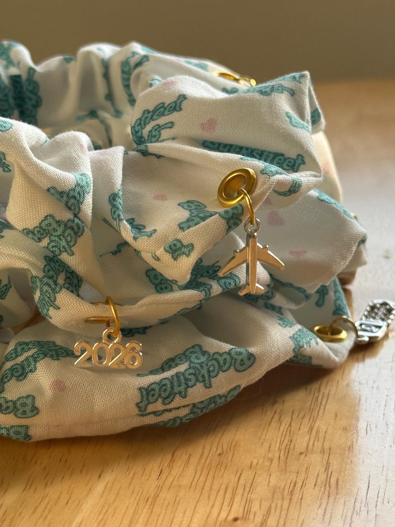 May include: A white scrunchie with a teal pattern and gold charms. The scrunchie has a repeating pattern of teal teddy bears and the word "Backstreet". Gold charms include a small airplane and the number "2026".