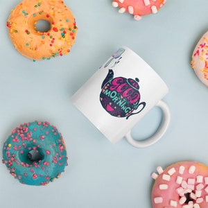 May include: White ceramic mug featuring a black and pink teapot design. The teapot has the words "Good Morning" written on it. The mug is surrounded by colourful doughnuts.