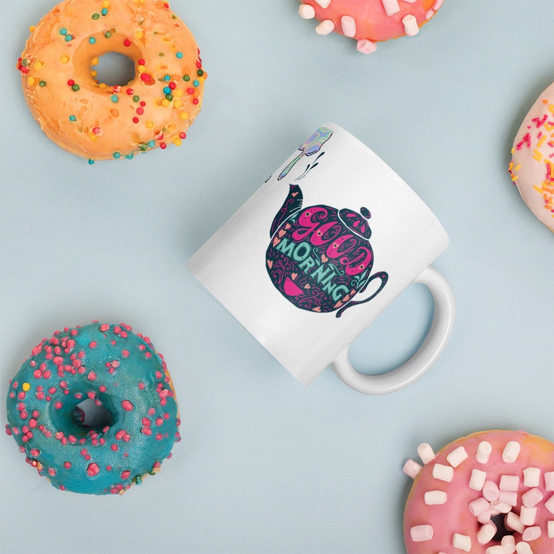 May include: White ceramic mug featuring a black and pink teapot design. The teapot has the words "Good Morning" written on it. The mug is surrounded by colourful doughnuts.