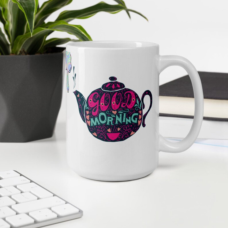 May include: White ceramic coffee mug featuring a black and white illustration of a teapot with the words "good morning" in pink and teal lettering.