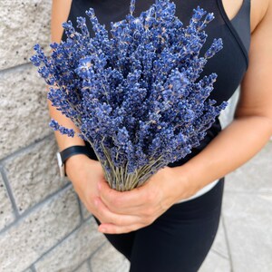 Puede incluir: Un ramo de flores de lavanda secas en tonos de morado y azul. Las flores est&aacute;n atadas con una cuerda natural y las sostiene una persona.