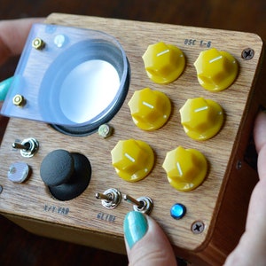 Könnte beinhalten: Eine Holzkiste mit einem Lautsprecher, sechs gelben Knöpfen mit der Beschriftung "OSC 1-6", zwei Kippschaltern und einem Joystick. Die Kiste hat eine blaue LED-Leuchte und den Text "X/Y VAR" und "GLIDE" auf der Vorderseite.