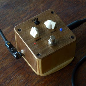 May include: A wooden guitar effects pedal with two knobs labelled "Shape" and "Depth", a toggle switch labelled "Shape", and a footswitch labelled "Build". The pedal has a blue LED light that is on.