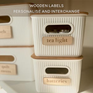 Könnte beinhalten: Weiße Aufbewahrungsboxen mit Holzdeckeln und Etiketten. Die Etiketten lauten "tea light" und "batteries". Die Boxen haben eine gerippte Textur und ovale Griffe. Der Text "WOODEN LABELS PERSONALIZE AND INTERCHANGE" steht oben.