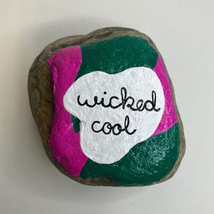 Peut inclure: Une pierre peinte avec les mots "wicked cool" en écriture noire sur un fond blanc en forme de nuage. La pierre est peinte avec des sections vertes et roses.