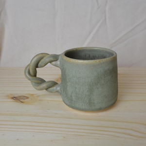 Może przedstawiać: Ręcznie robiony ceramiczny kubek w stonowanym zielonym odcieniu. Kubek ma unikalny uchwyt, który wygląda jak skręcona lina. Wnętrze i obręcz mają jaśniejszy, żółtawy odcień. Kubek stoi na jasnej drewnianej powierzchni.