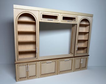 May include: A miniature wooden entertainment center with a natural finish. It features a central open space, two arched bookshelves, and lower cabinets with doors and drawers. The unit is designed for a dollhouse or craft project.