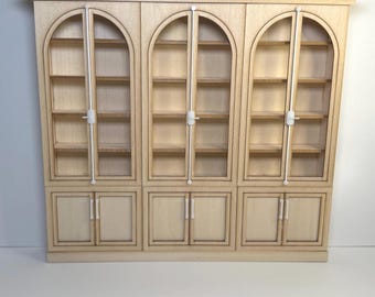 May include: A light-colored wooden cabinet with three arched glass-fronted sections above three solid-door sections. The cabinet has a simple, elegant design, with white hardware and a decorative top.