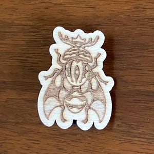 May include: A wooden laser-cut beetle-shaped ornament. The beetle has detailed wings and body features, with a light brown color and a white outline. The ornament is placed on a dark brown wooden surface.