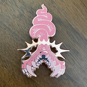 May include: A pink enamel pin with a rose gold outline. The pin features a stylized, abstract design with a layered, swirling top and a detailed, symmetrical base with starburst-like elements. The design incorporates shades of pink.