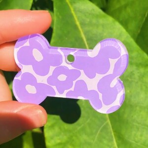 Puede incluir: Chapa para perro de color morado en forma de hueso con un patr&oacute;n floral blanco.