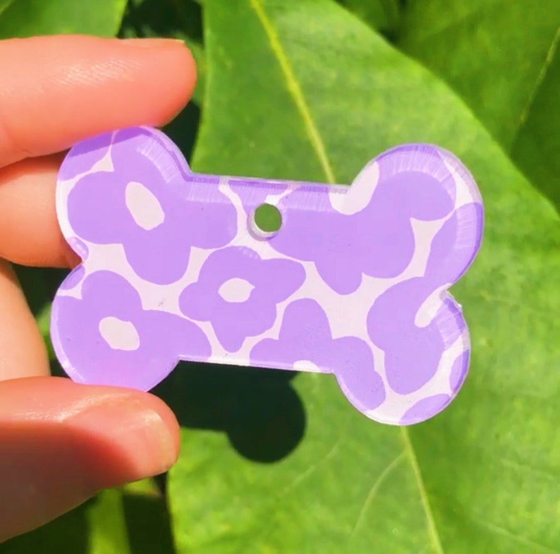Puede incluir: Chapa para perro de color morado en forma de hueso con un patr&oacute;n floral blanco.