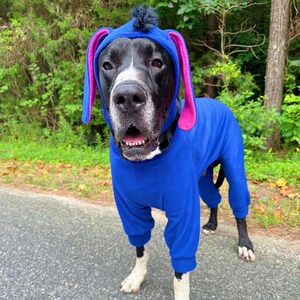 May include: A blue dog onesie featuring a hood with long, pink-lined ears and a small black tuft of hair on top. The garment covers the torso and legs, with ribbed cuffs at the ends of the sleeves and pant legs, designed for a canine to wear.