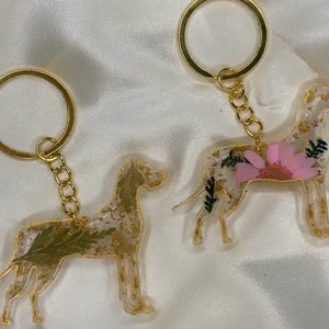 May include: Two gold keychains featuring resin dog silhouettes with pressed flowers and gold glitter. One dog is white with pink flowers and the other is white with green leaves.