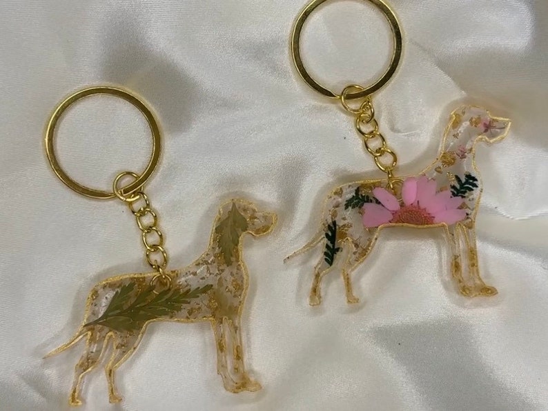 May include: Two gold keychains featuring resin dog silhouettes with pressed flowers and gold glitter. One dog is white with pink flowers and the other is white with green leaves.
