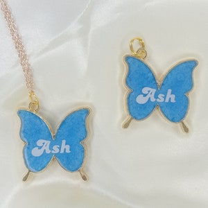 Puede incluir: Dos collares de mariposa de oro con alas azules y el nombre "Ash" escrito en blanco en cada uno.
