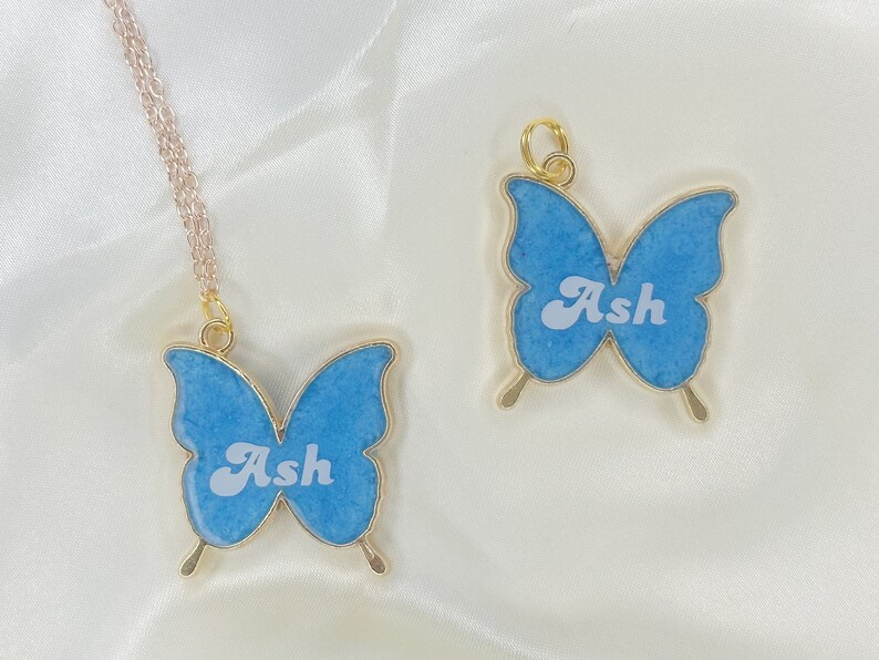 Puede incluir: Dos collares de mariposa de oro con alas azules y el nombre "Ash" escrito en blanco en cada uno.