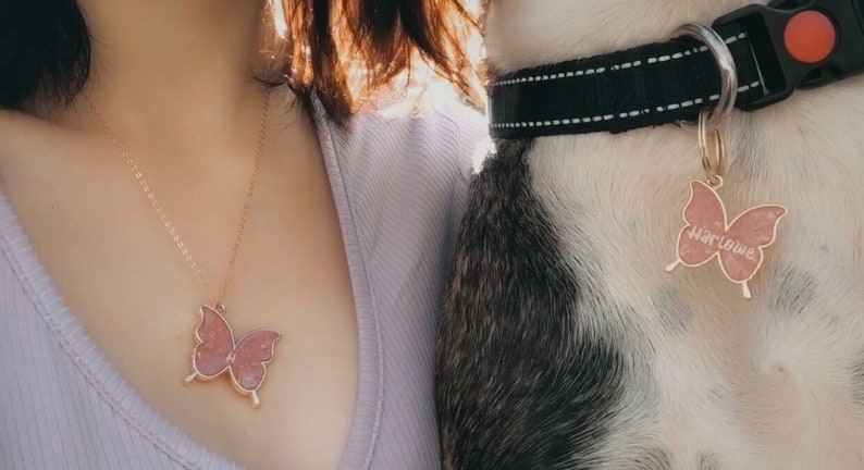 Puede incluir: Una persona lleva un collar de oro con un colgante de mariposa rosa. Un perro lleva un collar negro con una etiqueta de mariposa rosa que dice "Harlowe".