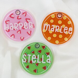 Puede incluir: Tres etiquetas de identificaci&oacute;n redondas y coloridas para mascotas con diferentes dise&ntilde;os de frutas. La etiqueta rosa tiene un dise&ntilde;o de sand&iacute;a y dice "Harper". La etiqueta naranja tiene un dise&ntilde;o de lim&oacute;n y dice "Marlee". La etiqueta verde tiene un dise&ntilde;o de sand&iacute;a y dice "Stella".