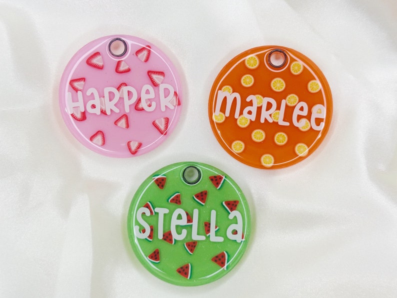 Puede incluir: Tres etiquetas de identificaci&oacute;n redondas y coloridas para mascotas con diferentes dise&ntilde;os de frutas. La etiqueta rosa tiene un dise&ntilde;o de sand&iacute;a y dice "Harper". La etiqueta naranja tiene un dise&ntilde;o de lim&oacute;n y dice "Marlee". La etiqueta verde tiene un dise&ntilde;o de sand&iacute;a y dice "Stella".