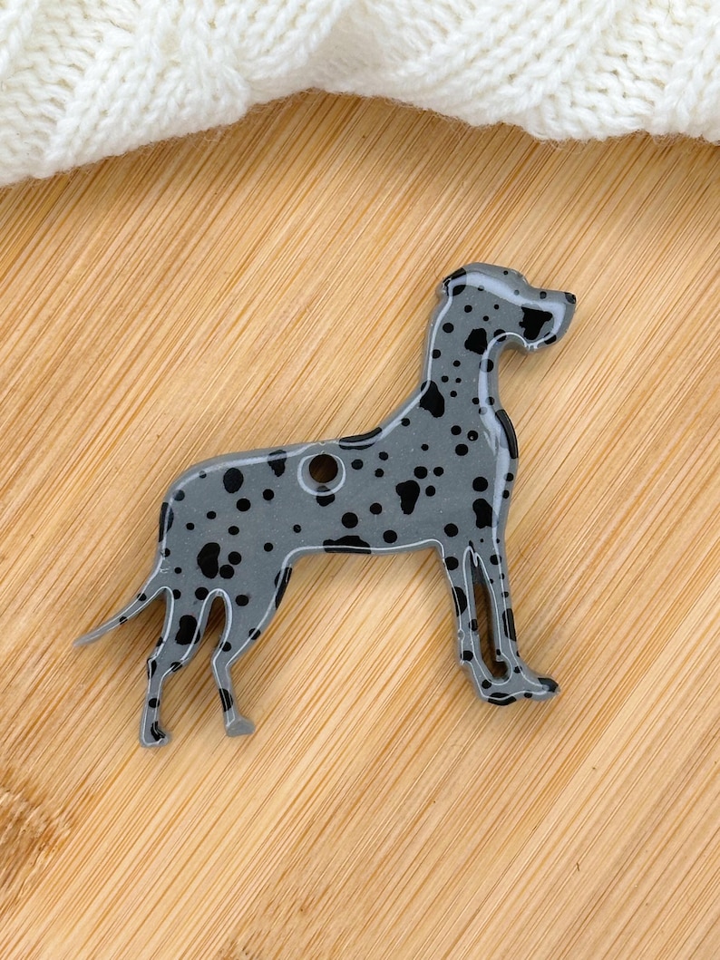 May include: A gray and black Great Dane dog shaped charm with black spots. The charm is made of resin and has a hole for hanging.