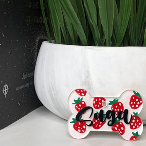 May include: White bone-shaped dog tag with a red and green strawberry pattern and the name "Suga" written in black.