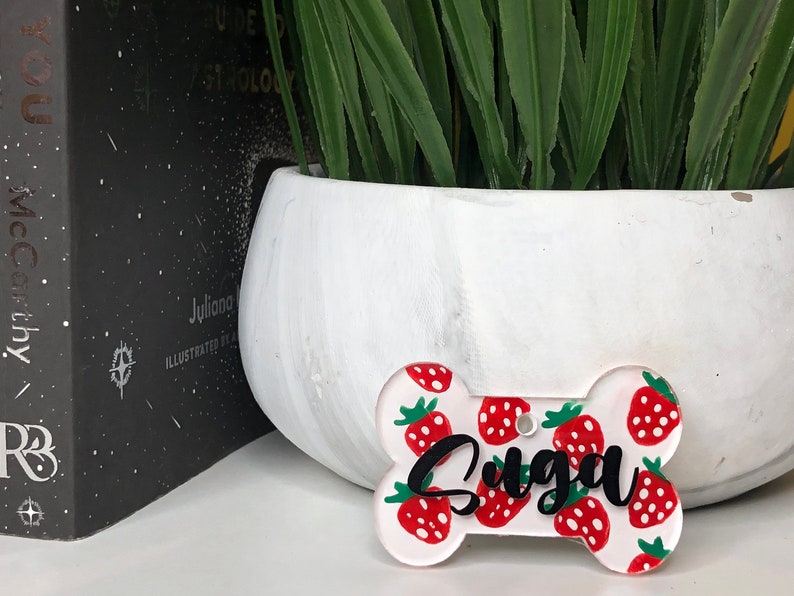 May include: White bone-shaped dog tag with a red and green strawberry pattern and the name "Suga" written in black.