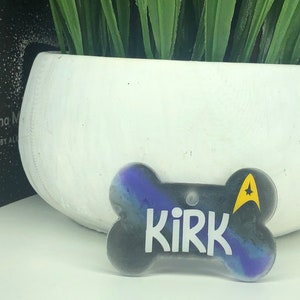 Puede incluir: Una placa de identificaci&oacute;n para perro en forma de hueso, de color negro y morado, con la palabra "Kirk" y un emblema amarillo de la Flota Estelar.