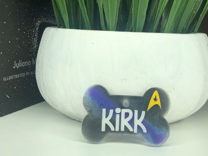 Puede incluir: Una placa de identificaci&oacute;n para perro en forma de hueso, de color negro y morado, con la palabra "Kirk" y un emblema amarillo de la Flota Estelar.
