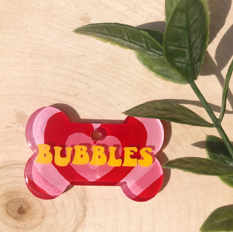 Puede incluir: Una etiqueta para perro con forma de hueso roja y rosa con la palabra "BUBBLES" en letras amarillas.