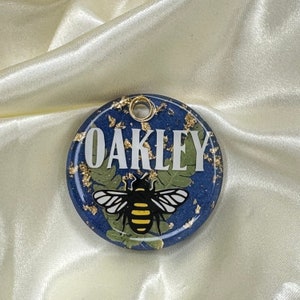 Puede incluir: Etiqueta redonda de resina azul con motas doradas, una abeja blanca con rayas amarillas y el nombre "OAKLEY" en letras blancas.