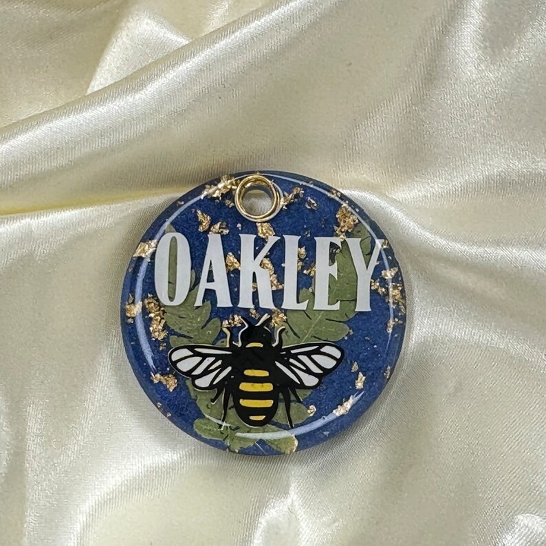 Puede incluir: Etiqueta redonda de resina azul con motas doradas, una abeja blanca con rayas amarillas y el nombre "OAKLEY" en letras blancas.