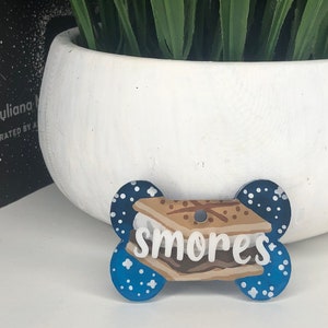 May include: Blue and white dog tag shaped like a bone with a white and brown s'more graphic and the word "smores" written in white.