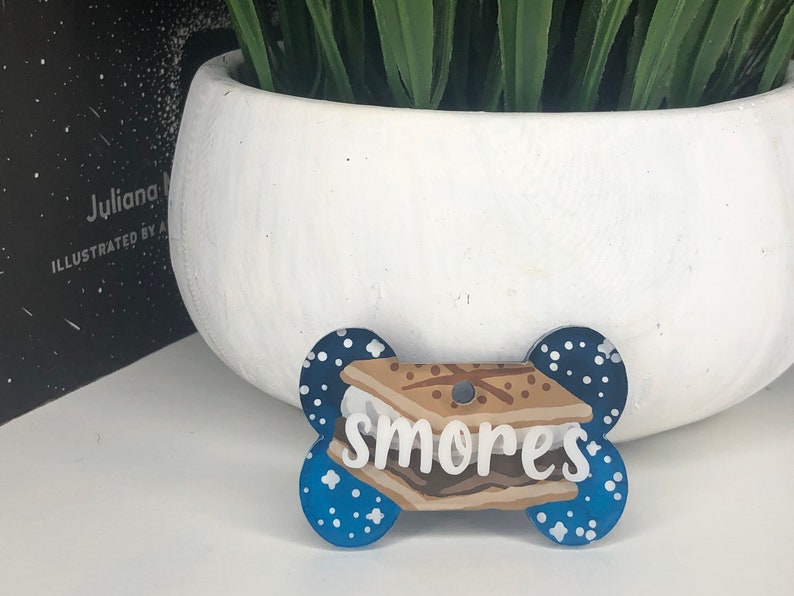 May include: Blue and white dog tag shaped like a bone with a white and brown s'more graphic and the word "smores" written in white.
